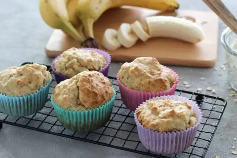 Fünf leckere Muffins ohne Zucker in bunten Förmchen, daneben Bananen und eine Schüssel Mehl.