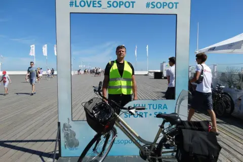 Mężczyzna w kamizelce odblaskowej pozuje przy rowerze na tle ramki z napisem #LOVES_SOPOT. Idealne miejsce na zdjęcie po zwiedzaniu tras rowerowych Sopot.