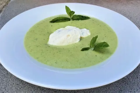 Velouté de courgette onctueux, garni d'une quenelle de fromage frais et de feuilles de menthe, servi dans une assiette blanche.