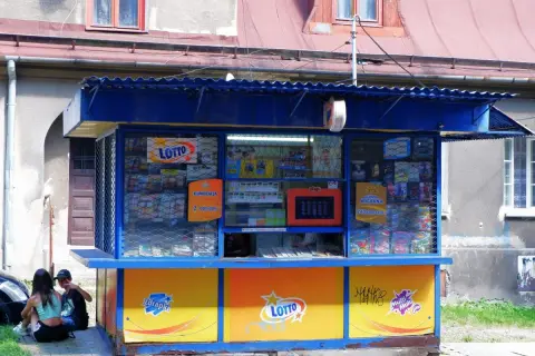 Kiosk Lotto z ofertą zdrapek. Czy warto kupować zdrapki na stacjach benzynowych czy lepiej w kolekturze? Dwoje młodych ludzi siedzi obok.