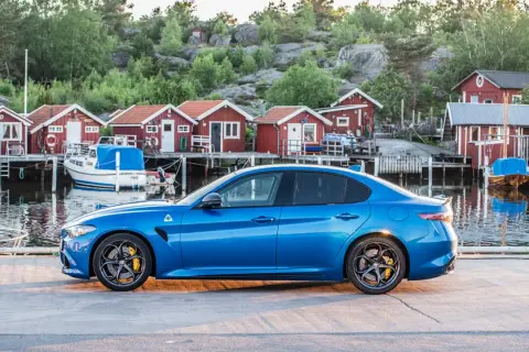 Blå sportbil vid en svensk hamn med röda sjöbodar. Undrar hur långt har bilen gått på denna vackra resa?