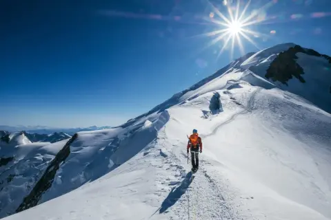 Un alpiniste solitaire progresse sur une crête enneigée, le soleil éclatant au-dessus du sommet du Mont-Blanc.
