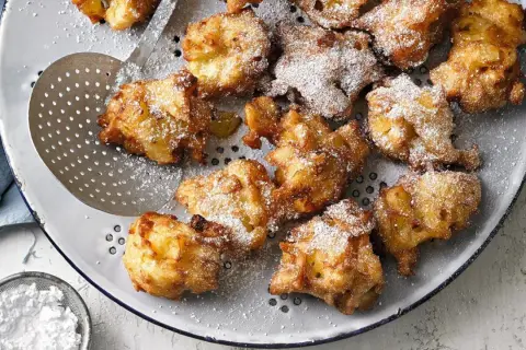 Goldbraune Apfelkrapfen, frisch aus der Pfanne, mit Puderzucker bestäubt, liegen auf einem Sieb.