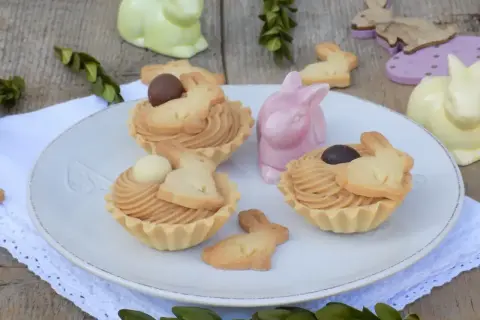 Wielkanocne babeczki z mascarpone, ozdobione ciasteczkami w kształcie króliczków i czekoladowymi jajeczkami, na talerzu wśród ceramicznych zajączków.