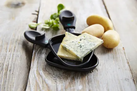 Tranches de bleu de Gex prêtes pour la raclette, avec des pommes de terre et de la verdure sur fond de bois.