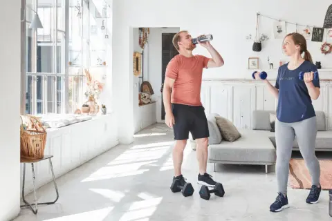 Un couple fait de l'exercice biceps maison avec des haltères bleus dans un salon lumineux.