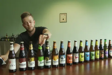 Mężczyzna wybiera piwo bez drożdży z rzędu butelek. Widać różne rodzaje piw, m.in. APA, Koźlak, Złote Lwy.