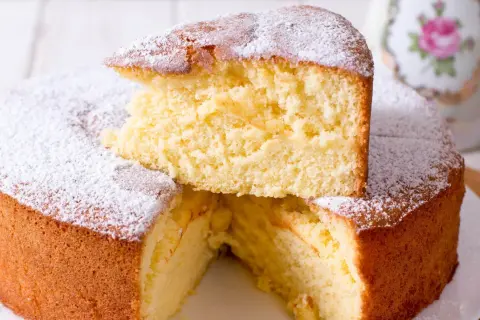 En fluffig vegansk sockerkaka, pudrad med florsocker. En bit är upplagd ovanpå resten av kakan.