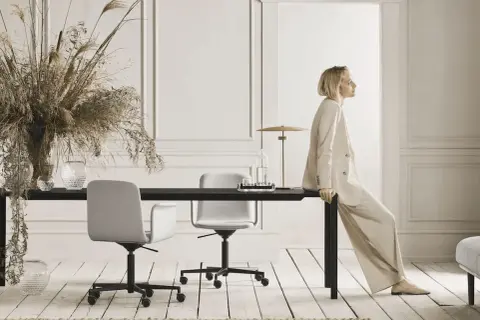 Eine Frau in einem beigen Anzug sitzt auf einem schwarzen Tisch. Der Raum ist in neutralen Farben gehalten und mit Trockenblumen und Glasvasen dekoriert.