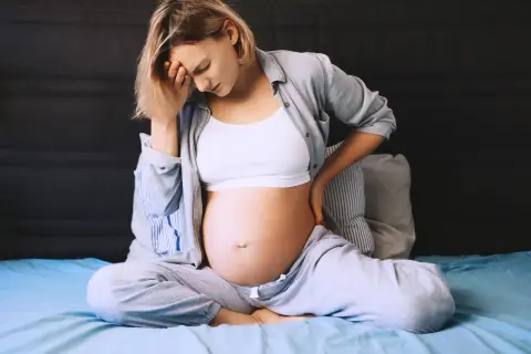 Ciężarna kobieta z bólem krocza w ciąży, trzymająca się za głowę i plecy, siedzi na łóżku.