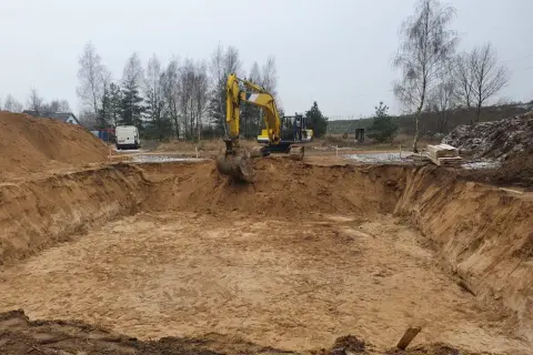 Koparka pracuje przy wykopy pod fundamenty. Widać głęboki dół, usypiska ziemi i materiały budowlane.
