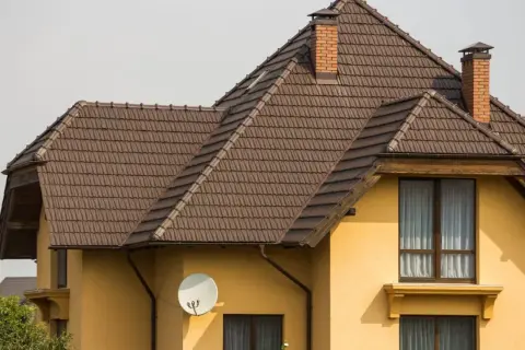 Dach naczółkowy konstrukcja z brązowej dachówki, z ceglanym kominem i anteną satelitarną na żółtej ścianie domu.