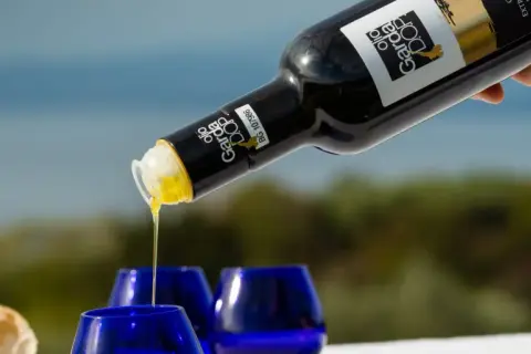 Versando olio Garda DOP in bicchierini blu, con pane fresco sullo sfondo.