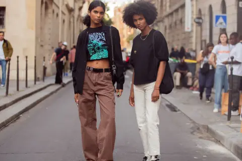 Zwei Frauen in lässiger Kleidung, eine mit brauner Cargohose und Crop-Top, die andere mit weißer Hose und schwarzem Shirt, stehen auf einer Straße.