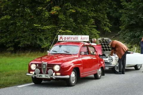 Röd Saab 96 V4 med "M-patrull" skylt på taket. En man tittar i motorhuven på en vit bil, en kvinna står bredvid.