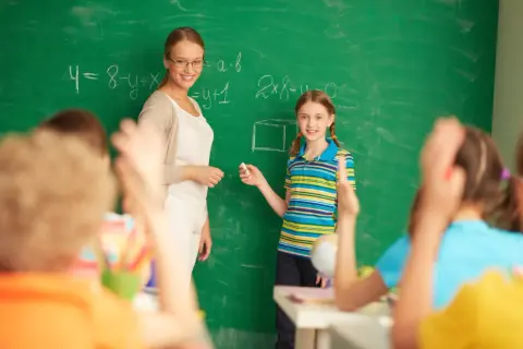 Nauczycielka dyplomowana z uśmiechem obserwuje uczennicę przy tablicy. To przykładowa ocena pracy nauczyciela.