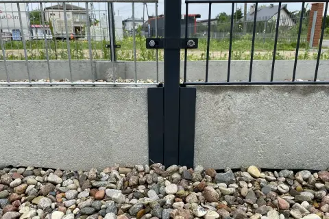 Metalowy łącznik słupka ogrodzenia do betonowej podmurówki. Kamienie ozdobne wokół.