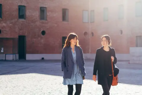 Las hermanas Arce caminan juntas bajo el sol, conversando animadamente frente a un edificio de ladrillo.