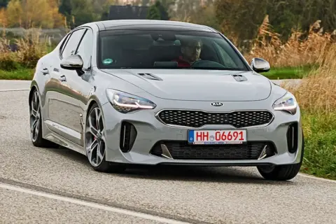 Szary Kia Stinger GT z kierowcą za kierownicą jedzie po asfaltowej drodze, otoczony suchą trawą i drzewami.