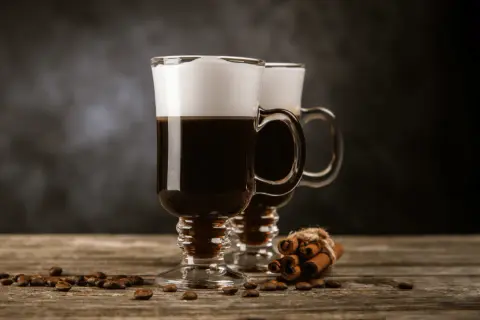Två glas med irish coffee, toppade med grädde, står på ett träbord. Kaffebönor och kanelstänger ligger utspridda.