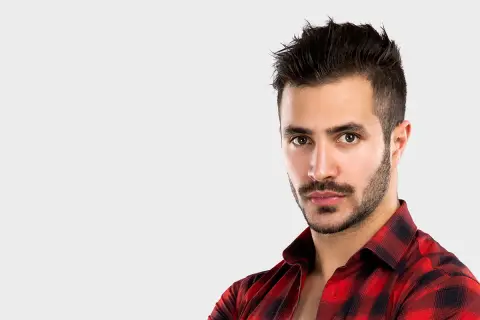 Un homme au regard intense, avec une barbe mouche bien taillée, porte une chemise à carreaux rouges et noirs.