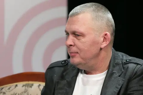 Adam z zespołu Raz Dwa Trzy w szarej marynarce, siedzi na kanapie.