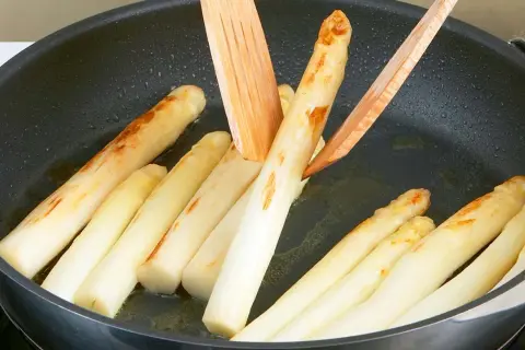Weißer Spargel brät in einer Pfanne mit etwas Öl. Holzspieße wenden die Stangen, die leicht gebräunte Stellen zeigen.