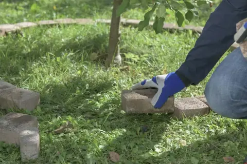 Ręce w rękawiczkach układają kostkę brukową, tworząc obrzeże wokół drzewka. Prosty sposób, jak zrobić obrzeże z kostki brukowej.
