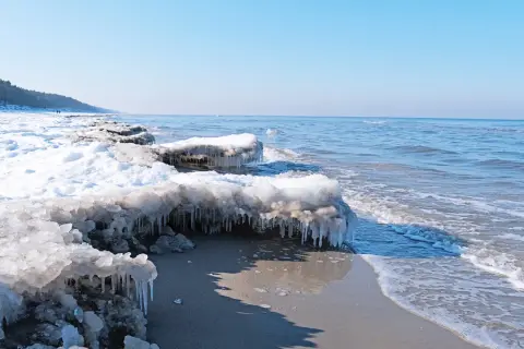 Lodowe sople zwisają z zamarzniętych kęp śniegu na plaży, gdy fale Bałtyku delikatnie obmywają brzeg. To widok z roku, kiedy zamarzł bałtyk rok.