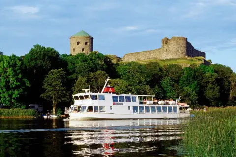 En vit båt från Strömma Boat Tours glider fram på vattnet, förbi grönskande träd och en gammal borg.