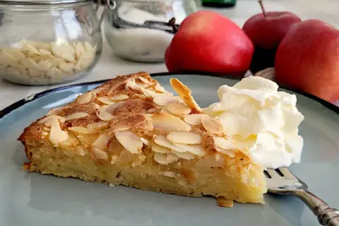 Ein Stück Apfelkuchen mit mandelkruste, garniert mit Sahne, auf einem blauen Teller. Im Hintergrund Äpfel und Mandelblättchen.