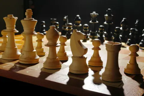 Białe figury szachowe, w tym król, hetman i goniec, ustawione na szachownicy. Ciemne figury w tle.
