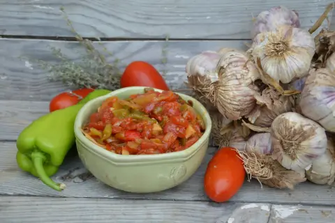 Un bol de piperade basque aux poivrons verts, tomates et oignons, avec des gousses d'ail et des herbes sur fond de bois.