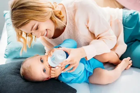Uśmiechnięta mama karmi butelką dziecko. Ile powinno trwać karmienie butelką? To zależy od malucha.