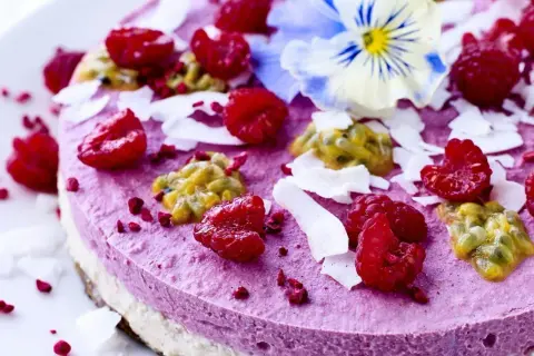 En vacker, rund kaka i rosa färg, dekorerad med hallon, passionsfrukt, kokosflingor och en blå viol.