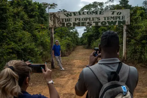 Un cartel de "Bienvenido al Templo del Pueblo Jonestown" se alza sobre un camino de tierra. Dos personas toman fotos, una de ellas posa cerca del cartel, un sombrío recordatorio de la masacre de Jonestown.