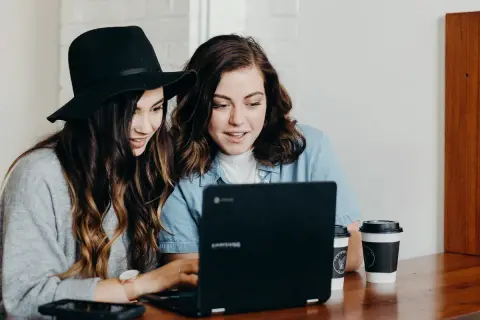 Två unga kvinnor diskuterar sin skuldkvot och bolån framför en laptop, med kaffekoppar på bordet.