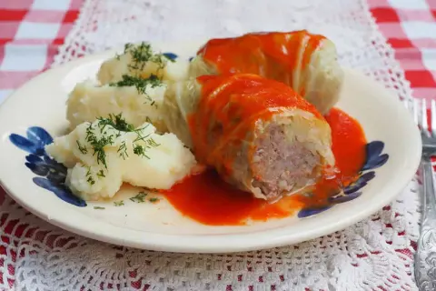 Gołąbki z kapusty włoskiej, podane z purée ziemniaczanym i polane sosem pomidorowym, ozdobione koperkiem.