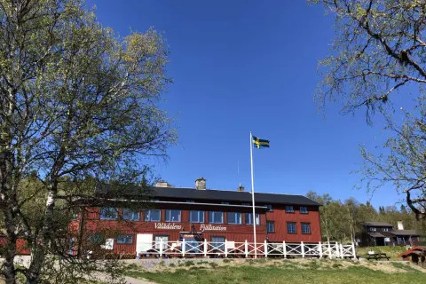 STF Vålådalen fjällstation i solsken med svensk flagga. Träd med gröna blad ramar in den röda byggnaden.