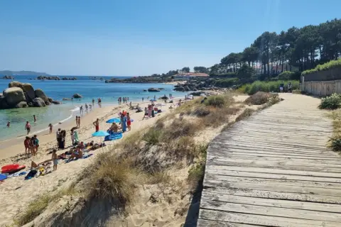 Gente disfrutando del sol y el mar en la playa Pedras Negras. Un camino de madera bordea la arena dorada y las dunas.