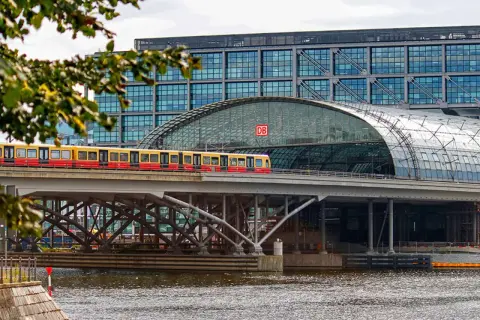 Ein roter und gelber Zug fährt über eine Brücke vor dem Berliner Hauptbahnhof. Die Verbindung Berlin Dresden ist hier gut sichtbar.