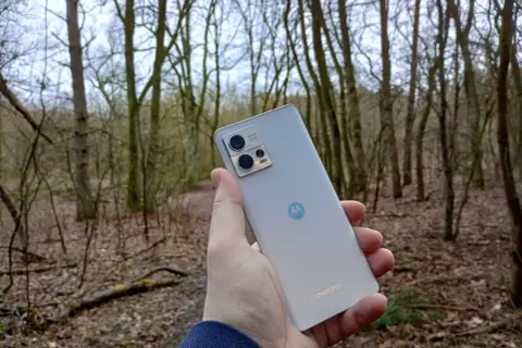 Dłoń trzyma srebrny smartfon Motorola G13 na tle lasu. Sprawdź motorola g13 dane techniczne, by poznać jego możliwości.