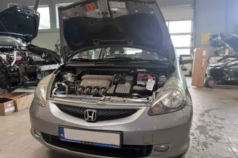 Srebrna Honda Jazz z otwartą maską, widoczny silnik i akumulator. Wymiana żarówki Honda Jazz w warsztacie.