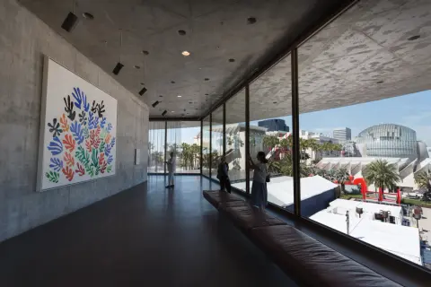 Inside a modern art museum, a colorful Matisse cutout hangs on a concrete wall. Visitors admire the art, enjoying the view of the city skyline.