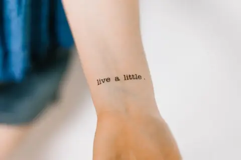 Ein Tattoo mit der Aufschrift "live a little" in Schreibmaschinenschrift auf einem Handgelenk.