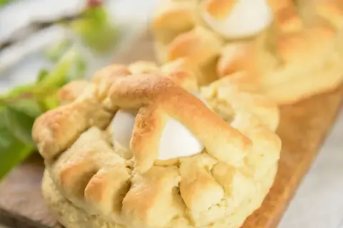 Dolci pasquali pugliesi con uova intere, decorati con impasto intrecciato, su un tagliere di legno.