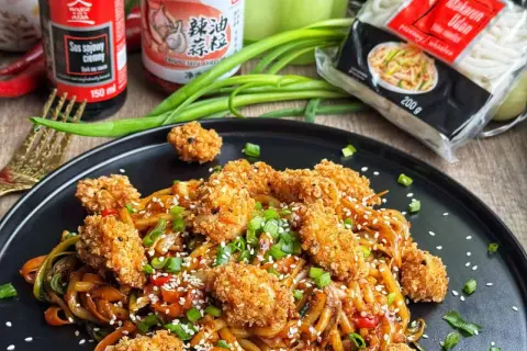 Pyszne udon z kurczakiem w panierce, posypane sezamem i szczypiorkiem. Obok sos sojowy i opakowanie makaronu udon.