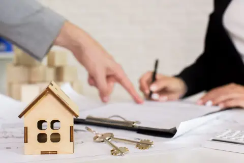 Un perito per valutazione casa indica un documento mentre un'altra persona firma. Chiavi e modellino di casa sul tavolo.