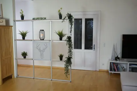 Ein Regal mit Pflanzen und Deko hilft, den Raum optisch zu trennen. Eine Tür und ein Fernseher sind im Hintergrund zu sehen.