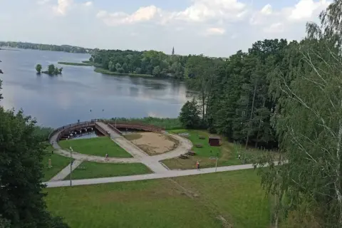 Malowniczy widok na jezioro i promenadę w Szczecinku. Widać ścieżki idealne na rowerowy spacer, mostek i zieleń.